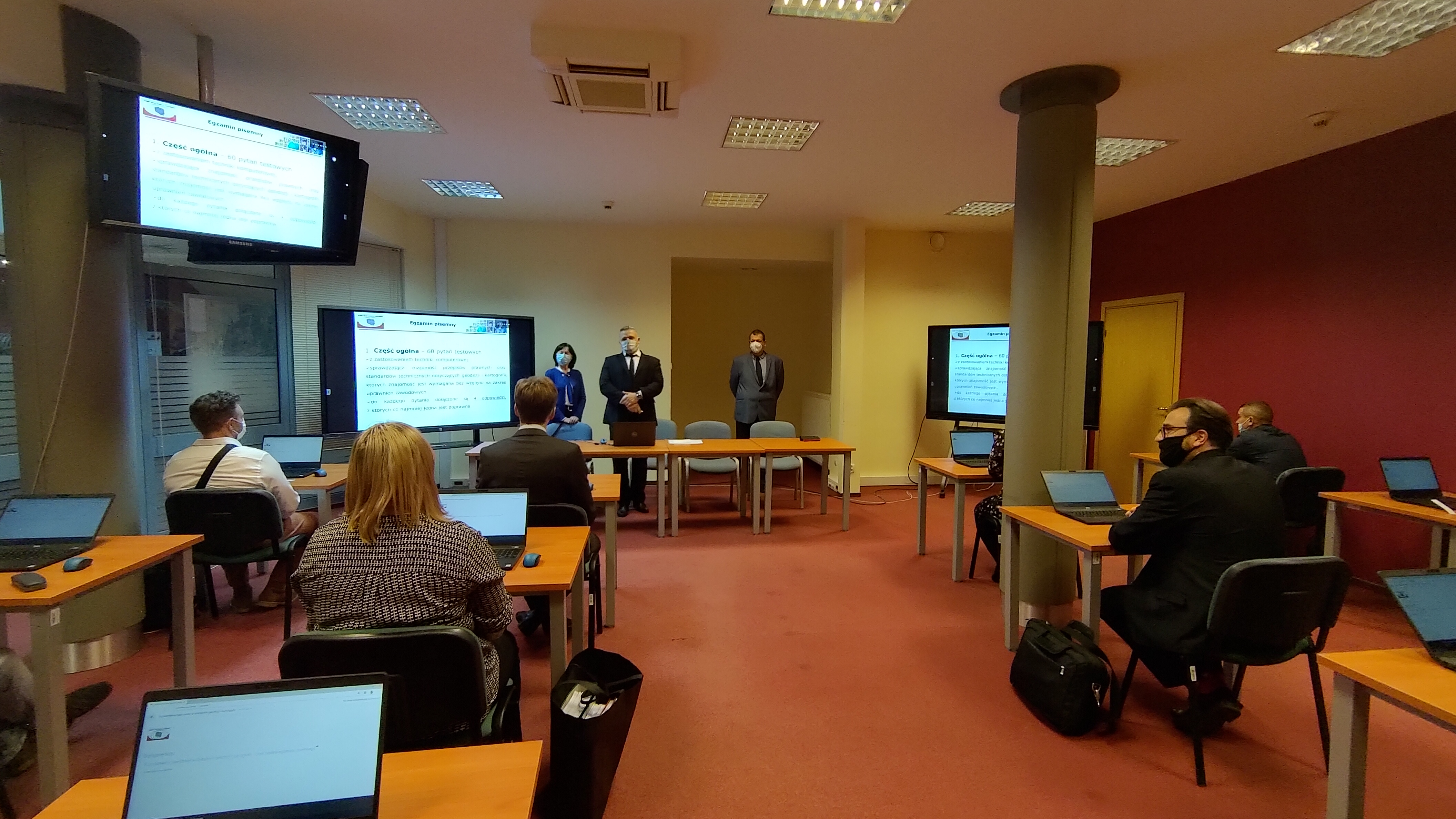 Zdjęcie przedstawia salę egzaminacyjną z członkami Komisji egzaminacyjnej i osobami przystępującymi do egzaminu siedzącymi pojedynczo przed laptopami