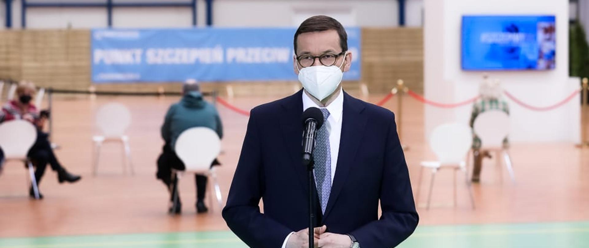 Premier Mateusz Morawiecki podczas brieifngu nt. szczepień. Premier stoi w hali w Skierniewicach, która została przerobiona na punkt szczepień. W tle osoby siedzące na krzesełkach i banery informujące o szczepieniach.