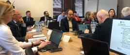 Kilkanaście osób siedzi na sali konferencyjnej. Na biurkach leżą laptopy.