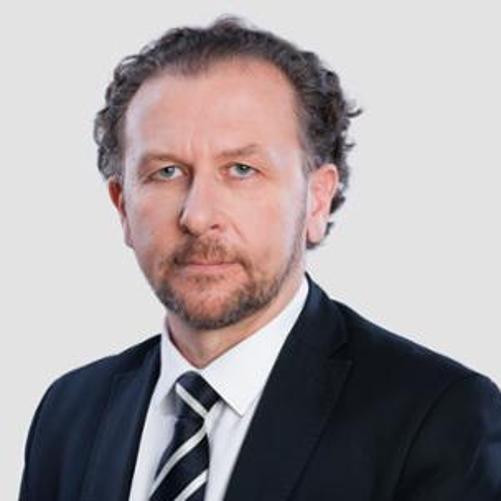 Na tle jasnoszarej ściany stoi dyrektor Bartold w czarnym garniturze.